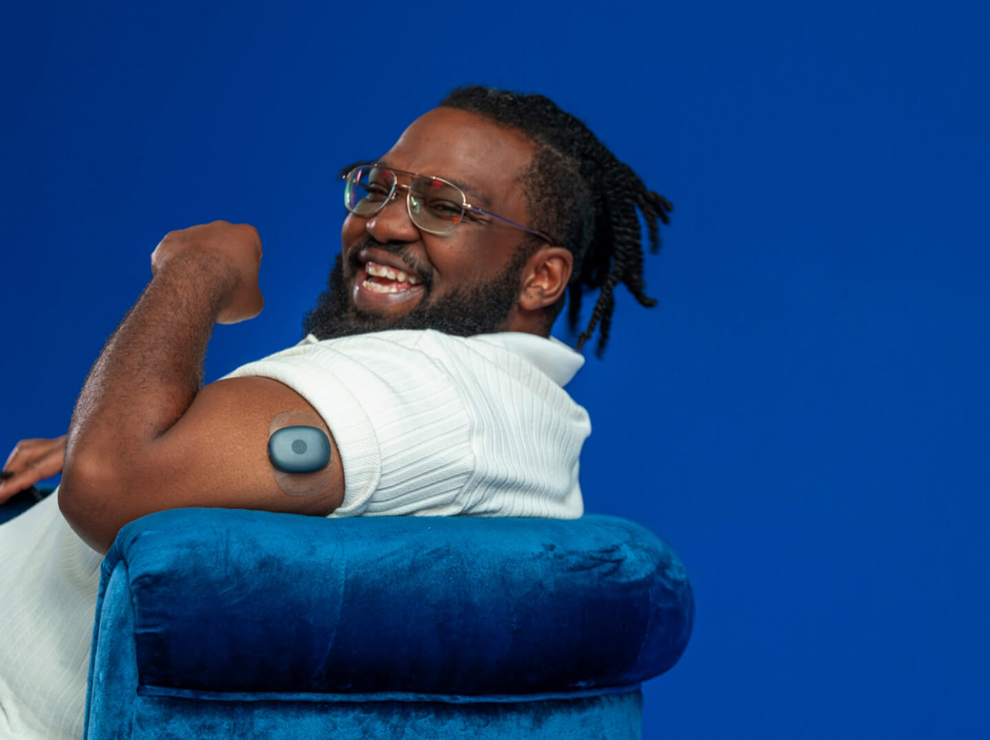 Eversense user smiling, flexing arm with a CGM sensor device attached, seated on a blue chair.