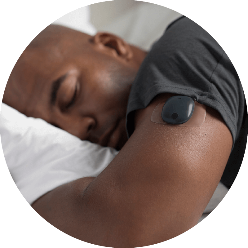 Person lies asleep on a pillow, wearing a small black CGM device on the upper arm for monitoring.
